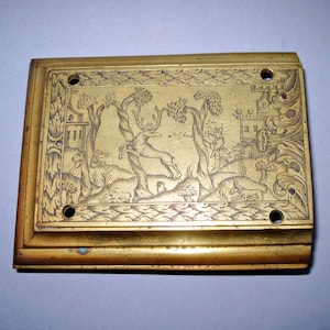May include: A rectangular brass plate with an engraved hunting scene. The scene depicts a deer leaping, a hunter, and a dog, set amidst trees and buildings. The plate has four holes and a raised border.