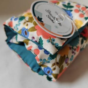 Könnte beinhalten: Ein weißer Stoffbeutel mit tealblauem Besatz und einem Blumenmuster. Der Beutel hat ein weißes Etikett, auf dem "Bouillotte Seche" mit einer kleinen Blumenillustration steht.
