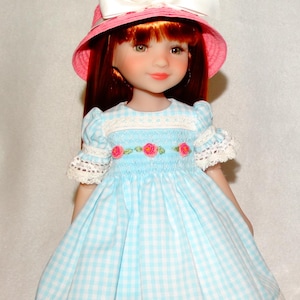 May include: A doll with red hair wearing a pink sun hat with a white bow and floral accents. The doll is dressed in a light blue and white gingham dress with lace trim and embroidered pink roses.