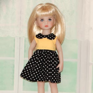 May include: A doll with blonde hair and blue eyes wearing a yellow top with a black polka dot collar and a black skirt with white polka dots. The doll is standing in front of a window.