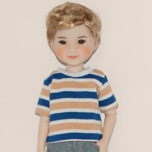 May include: A doll with blonde hair and freckles wearing a blue, tan, and white striped short-sleeved shirt and gray pants. The doll has rosy cheeks and is looking forward.