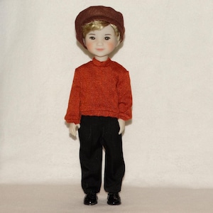 May include: A doll dressed in a rust-colored sweater, black pants, and black shoes. The doll is wearing a brown newsboy cap. The doll has blonde hair and a fair complexion. The doll is standing in front of a white background.
