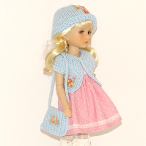 Puede incluir: Una muñeca con un gorro de punto azul claro, un cárdigan a juego de manga corta y un bolso, todos adornados con bordados florales. La muñeca viste un vestido rosa con lunares blancos y ribete de encaje blanco.