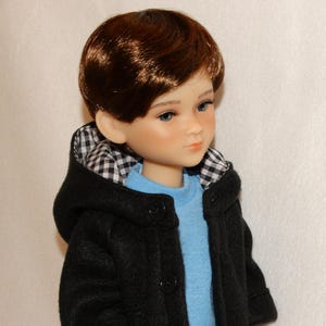 May include: A doll with brown hair and blue eyes, wearing a black coat with a gingham-lined hood over a blue sweater. The doll has a fair complexion and is looking slightly to the side.