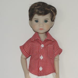 May include: A doll with brown hair and blue eyes wearing a short-sleeved button-down shirt with a red and white gingham check pattern. The shirt features a small chest pocket, a classic collar, and three white buttons down the center front.