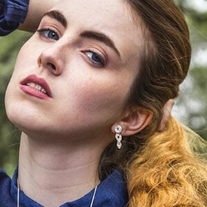May include: A woman with long, wavy, auburn hair is wearing a dark blue collared shirt with a silver necklace. She is holding her hair back with her right hand.