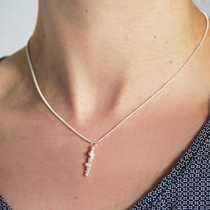 Puede incluir: Un collar de plata con un pequeño colgante hecho de varias cuentas de plata. El collar está puesto en el cuello de una persona.