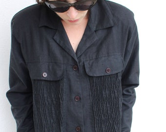 May include: A black long-sleeved shirt with a button-up front and a pleated detail on the front. The shirt has a collar and two pockets.