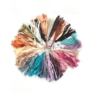 May include: A circular arrangement of multiple fabric tassels in a variety of colors including black, white, brown, tan, teal, pink, purple, and green. Each tassel is constructed from thin, frayed strips of fabric attached to a thin cord loop.