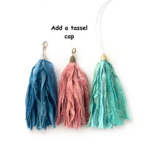 May include: Three fabric tassels in blue, pink, and turquoise, each featuring a metal cap at the top. The blue tassel has a silver lobster clasp, the pink has a bronze loop, and the turquoise has a gold cap with a white string. Text reads: Add a tassel cap.