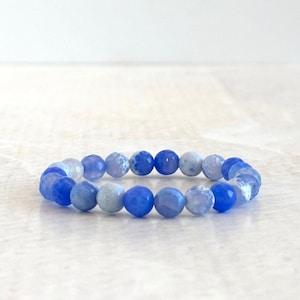 Puede incluir: Una pulsera elástica compuesta por cuentas redondas facetadas en varios tonos de azul, que van desde el azul cielo claro hasta el azul real intenso. Las cuentas tienen un aspecto semitranslúcido con sutiles vetas internas y patrones nubosos.