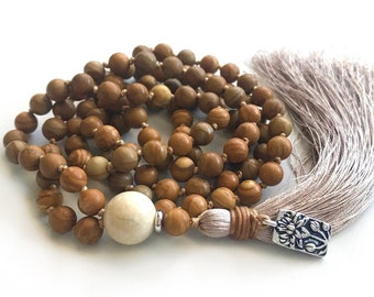 May include: A beaded necklace featuring round brown wooden beads, a single large off-white spherical bead, and a long light beige tassel. The tassel is attached to a silver-toned rectangular metal charm with an embossed floral pattern.