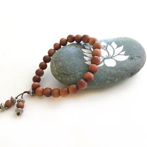 May include: A bracelet composed of smooth, round, medium-brown wooden beads strung on a dark cord. The bracelet features two dangling ends, each finished with a smaller wooden bead and a decorative, intricate silver-toned metal spacer bead.
