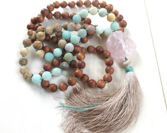 May include: A beaded necklace featuring a mix of smooth brown wooden beads, matte light blue amazonite beads, and patterned beige jasper beads. A large, raw, translucent pink rose quartz stone is attached to a long, shimmering beige tassel.