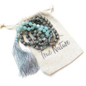 May include: A beaded necklace featuring round grey marbled beads and smooth light blue beads, finished with a long silver-colored tassel. The necklace rests on a beige drawstring pouch made of fabric with the text True Nature printed in grey.
