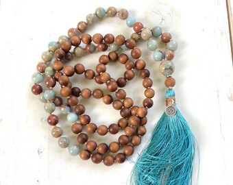 May include: A beaded necklace featuring a combination of smooth brown wooden beads and variegated blue and brown stone beads. The necklace is finished with a long, vibrant turquoise tassel attached to a small silver charm with an Om symbol.