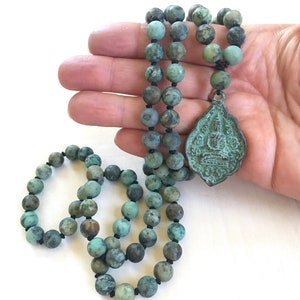 May include: A necklace featuring round, mottled turquoise-colored beads strung on black cord, accented with a central, shield-shaped metal pendant displaying a relief of a seated figure. The beads show natural variations in green and blue tones.