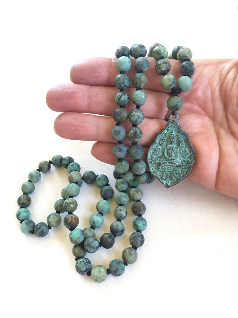 May include: A necklace featuring round, mottled turquoise-colored beads strung on black cord, accented with a central, shield-shaped metal pendant displaying a relief of a seated figure. The beads show natural variations in green and blue tones.