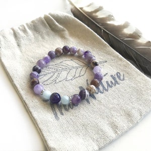 May include: A stretch bracelet composed of round beads in shades of purple, brown, and light blue, accented with small silver-toned metal spacers. The bracelet rests on a beige fabric pouch featuring a printed feather graphic and text.