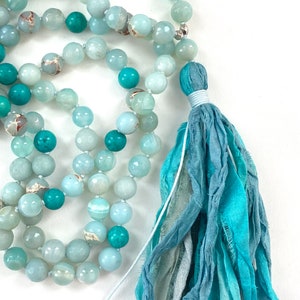 May include: A Throat Chakra Mala necklace featuring round, polished beads in shades of light blue, turquoise, and teal, accented with metallic spacers. The strand ends in a multi-layered, frayed fabric tassel in varying blue and teal tones.
