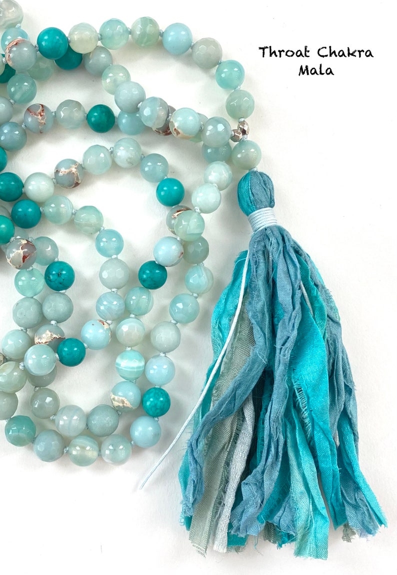 May include: A Throat Chakra Mala necklace featuring round, polished beads in shades of light blue, turquoise, and teal, accented with metallic spacers. The strand ends in a multi-layered, frayed fabric tassel in varying blue and teal tones.