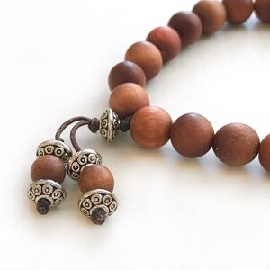 May include: A bracelet composed of smooth, round, medium-brown wooden beads strung on a dark cord. The bracelet features two dangling ends, each finished with a smaller wooden bead and a decorative, etched silver-toned metal spacer bead.