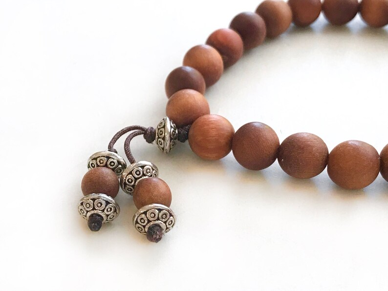 May include: A bracelet composed of smooth, round, medium-brown wooden beads strung on a dark cord. The bracelet features two dangling ends, each finished with a smaller wooden bead and a decorative, etched silver-toned metal spacer bead.