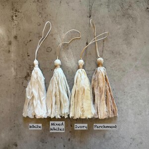 May include: Four fabric tassels with hanging loops, labeled White, Mixed Whites, Ivory, and Parchment. Each tassel features frayed edges and a tied top section. The colors range from bright white to a warm, light brown parchment tone.