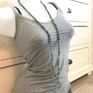 May include: A long necklace featuring 8mm knotted green and grey stone beads with a teardrop-shaped pendant. The text 8mm knotted Mala is visible in the upper left corner. The necklace is displayed on a grey tank top and denim shorts.