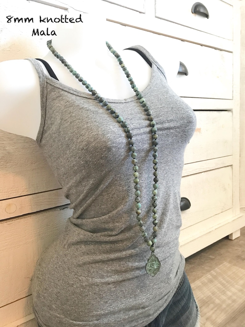 May include: A long necklace featuring 8mm knotted green and grey stone beads with a teardrop-shaped pendant. The text 8mm knotted Mala is visible in the upper left corner. The necklace is displayed on a grey tank top and denim shorts.