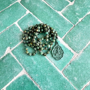 May include: A necklace featuring round, mottled green and brown beads strung on a dark cord, finished with a teardrop-shaped metal pendant that displays an embossed figure of a seated person in a meditative pose with intricate decorative borders.