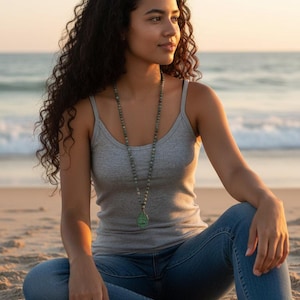 May include: A long beaded necklace featuring small, round, mottled green and grey stones strung together, ending in a teardrop-shaped green pendant with a textured surface, worn over a simple grey tank top and blue denim jeans.