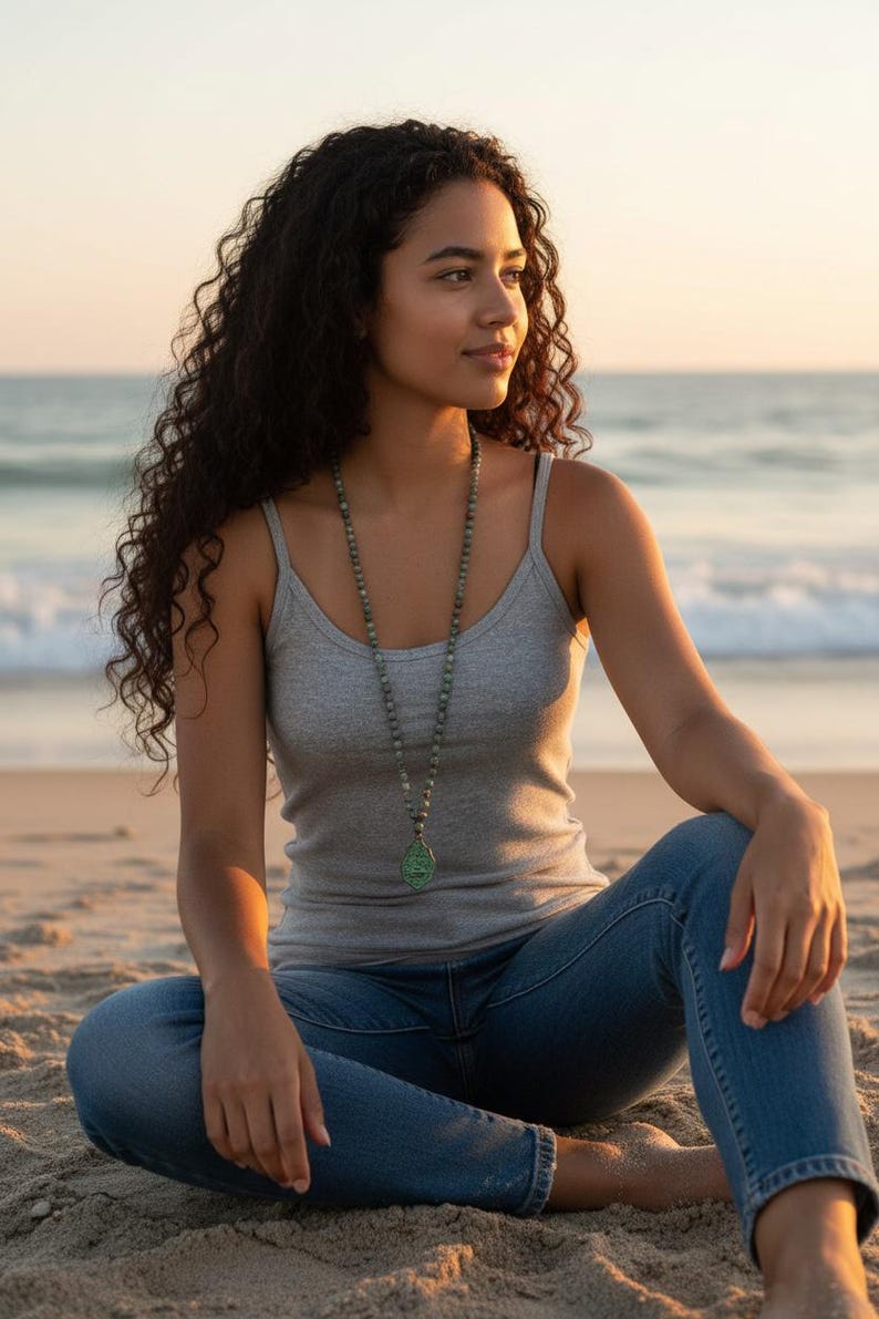 May include: A long beaded necklace featuring small, round, mottled green and grey stones strung together, ending in a teardrop-shaped green pendant with a textured surface, worn over a simple grey tank top and blue denim jeans.