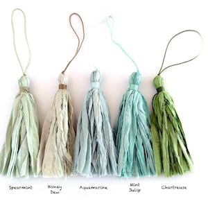 May include: Five fabric tassels with hanging loops, labeled Spearmint, Honey Dew, Aquamarine, Mint Julip, and Chartreuse. Each tassel is made of frayed, textured strips of cloth in varying shades of light green, beige, and blue-green.