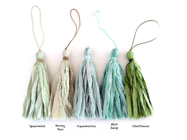 May include: Five fabric tassels with hanging loops, labeled Spearmint, Honey Dew, Aquamarine, Mint Julip, and Chartreuse. Each tassel is made of frayed, textured strips of cloth in varying shades of light green, beige, and blue-green.