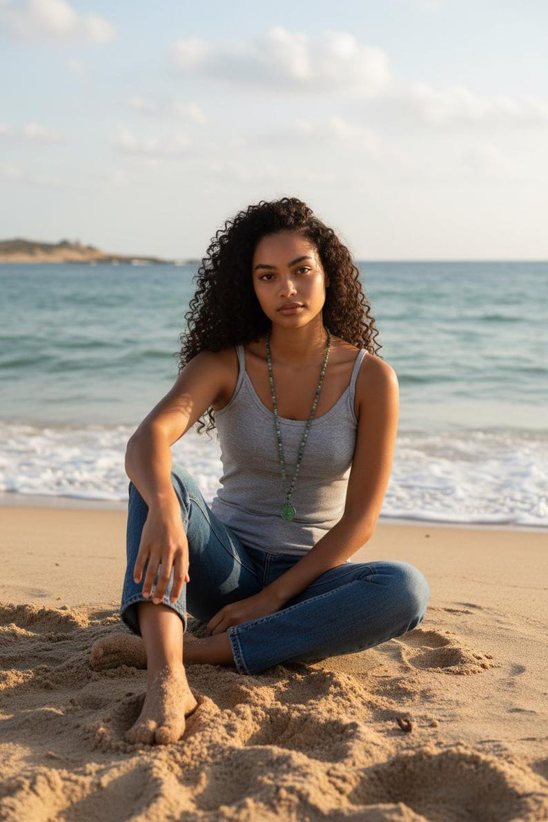 May include: A grey ribbed tank top, blue denim jeans, and a long beaded necklace featuring a green pendant. The necklace consists of small, multi-colored beads strung together, leading to a smooth, circular green stone pendant resting on the chest.