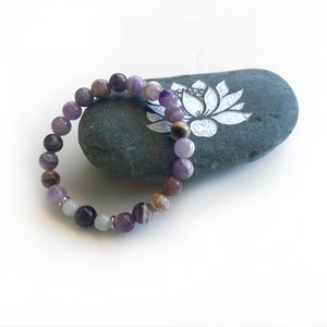 May include: A stretch bracelet composed of round beads in shades of purple, brown, and white, featuring smooth polished surfaces and interspersed with small silver-toned metal spacers, arranged in a circular loop on a white background.