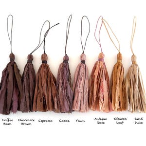 May include: Eight fabric tassels arranged in a row, each with a thin hanging loop. The colors range from dark brown to light beige, labeled as Coffee Bean, Chocolate Brown, Espresso, Cocoa, Fawn, Antique Rose, Tobacco Leaf, and Sand Dune.