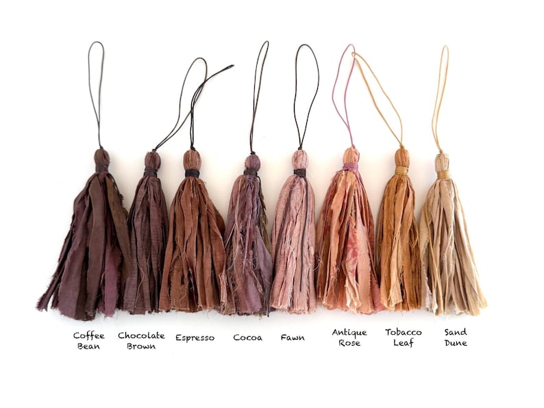 May include: Eight fabric tassels arranged in a row, each with a thin hanging loop. The colors range from dark brown to light beige, labeled as Coffee Bean, Chocolate Brown, Espresso, Cocoa, Fawn, Antique Rose, Tobacco Leaf, and Sand Dune.