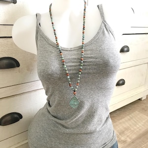 May include: A 6mm knotted Mala necklace featuring multi-colored stone beads in shades of teal, grey, and terracotta. The necklace includes a central pendant with a teal-toned, ornate, shield-shaped metal charm featuring an embossed design.