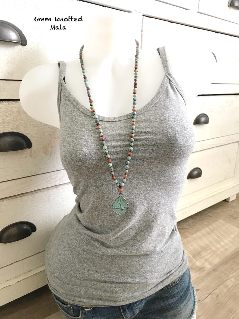 May include: A 6mm knotted Mala necklace featuring multi-colored stone beads in shades of teal, grey, and terracotta. The necklace includes a central pendant with a teal-toned, ornate, shield-shaped metal charm featuring an embossed design.