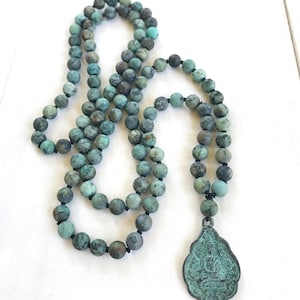 May include: A long necklace featuring round, matte-finish beads in shades of teal, turquoise, and dark gray, strung on a dark cord. The necklace includes a teardrop-shaped metal pendant with a weathered, oxidized green patina and a relief design.