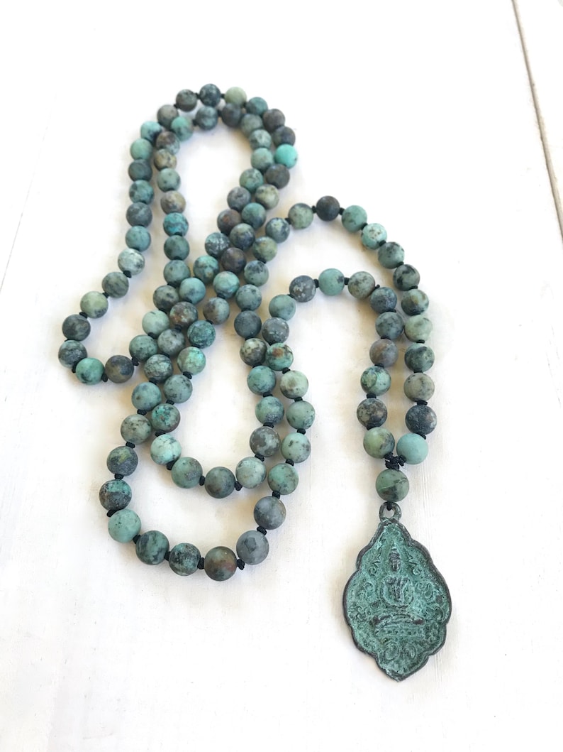 May include: A long necklace featuring round, matte-finish beads in shades of teal, turquoise, and dark gray, strung on a dark cord. The necklace includes a teardrop-shaped metal pendant with a weathered, oxidized green patina and a relief design.