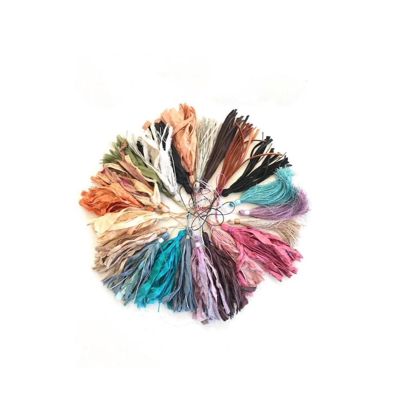 May include: A circular arrangement of multiple fabric tassels in a variety of colors including shades of pink, blue, green, brown, orange, and cream. Each tassel is constructed from thin, frayed strips of fabric attached to a thin cord loop.