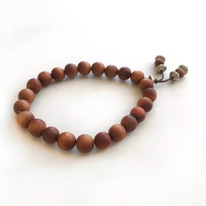 May include: A bracelet composed of smooth, round, medium-brown wooden beads strung on a cord. The ends of the cord feature two small, silver-toned metal beads with textured surfaces, providing a decorative finish to the circular beaded accessory.