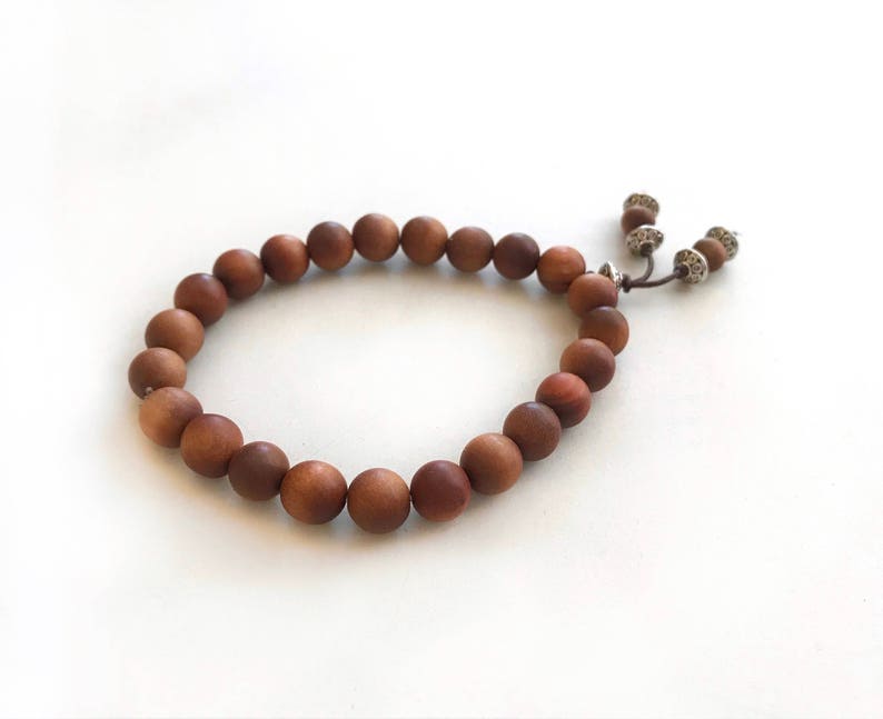 May include: A bracelet composed of smooth, round, medium-brown wooden beads strung on a cord. The ends of the cord feature two small, silver-toned metal beads with textured surfaces, providing a decorative finish to the circular beaded accessory.