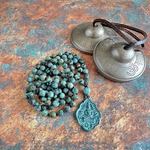 May include: A strand of round, mottled turquoise-colored beads with a dark, weathered pendant featuring a seated figure, paired with two circular bronze-toned metal cymbals connected by a dark brown leather cord, each engraved with Sanskrit text.
