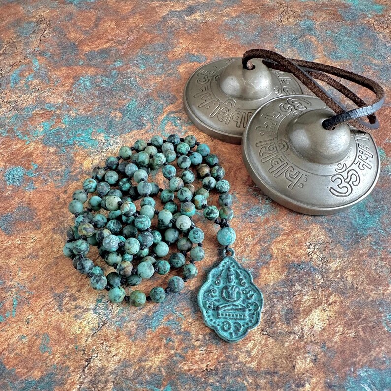 May include: A strand of round, mottled turquoise-colored beads with a dark, weathered pendant featuring a seated figure, paired with two circular bronze-toned metal cymbals connected by a dark brown leather cord, each engraved with Sanskrit text.