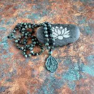 May include: A necklace featuring round, mottled green and black beads strung on a dark cord, ending in a metallic, shield-shaped pendant embossed with a seated figure. The necklace rests beside a smooth, dark gray stone with a white lotus design.