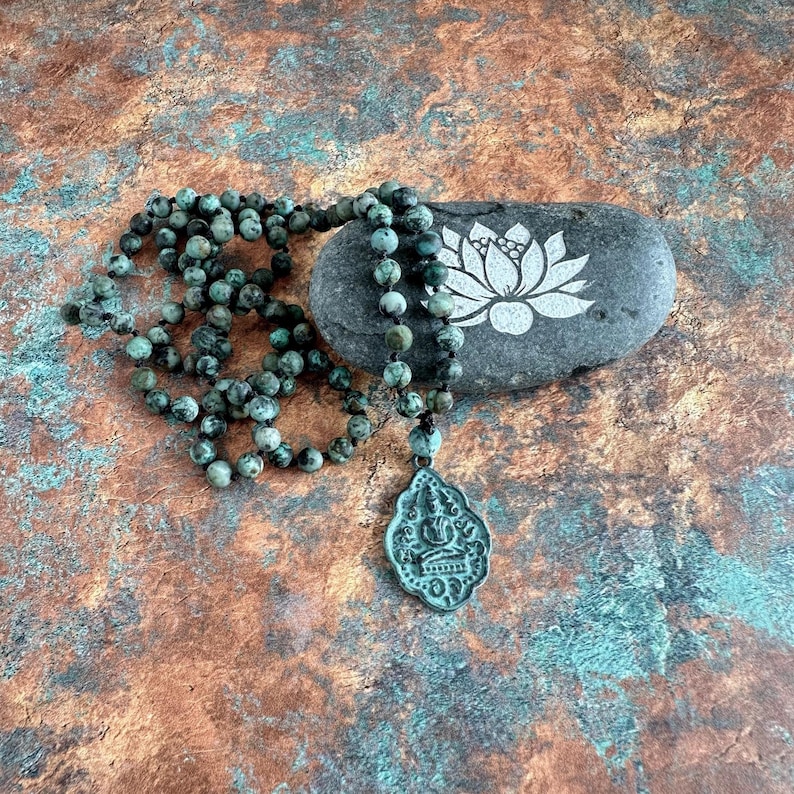 May include: A necklace featuring round, mottled green and black beads strung on a dark cord, ending in a metallic, shield-shaped pendant embossed with a seated figure. The necklace rests beside a smooth, dark gray stone with a white lotus design.